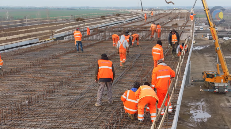 Constructori autostrăzi lucrând pe șantierul autostrăzii A0, cu utilaje și structuri în construcție, evidențiind progresul lucrărilor în noiembrie 2025. | sursa foto: Economica
